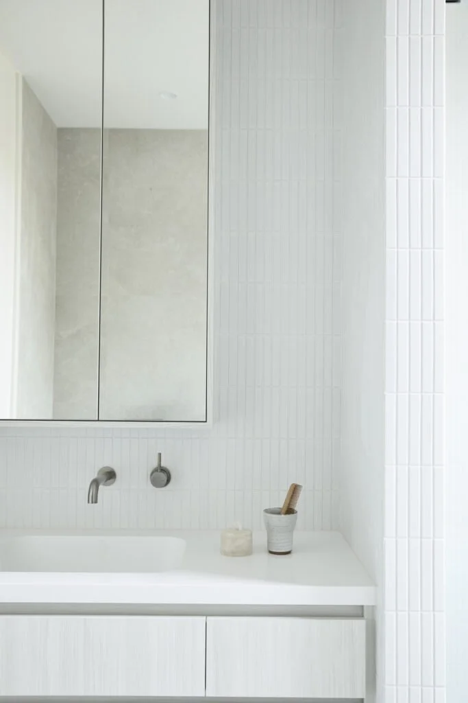 Minimalist bathroom sink area with white countertop, wall-mounted faucet, mirror, and small container with a toothbrush and dental floss, in a bright and modern space.