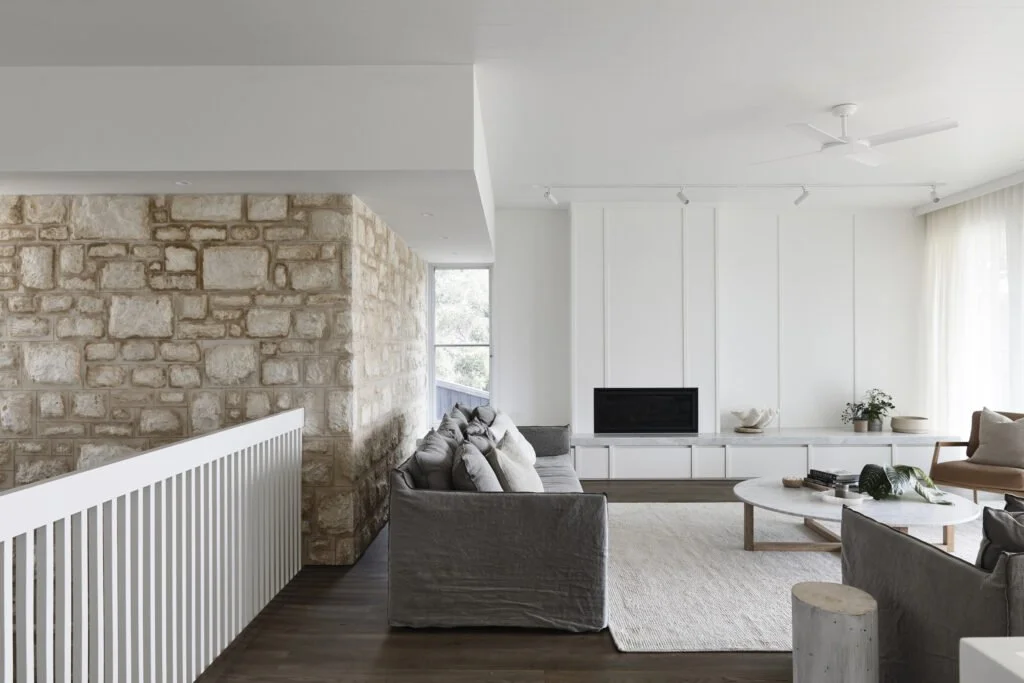 Modern living room with stone accent wall, gray sofa, white built-in fireplace, round coffee table, and large windows with curtains.