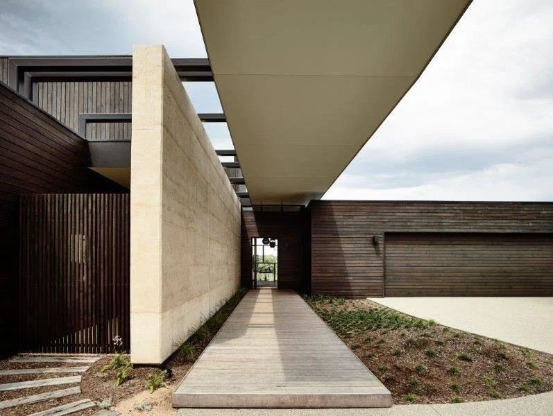 Modern architectural building with concrete, wood, and metal elements, featuring a pathway leading to a glass door, and a large overhanging roof.