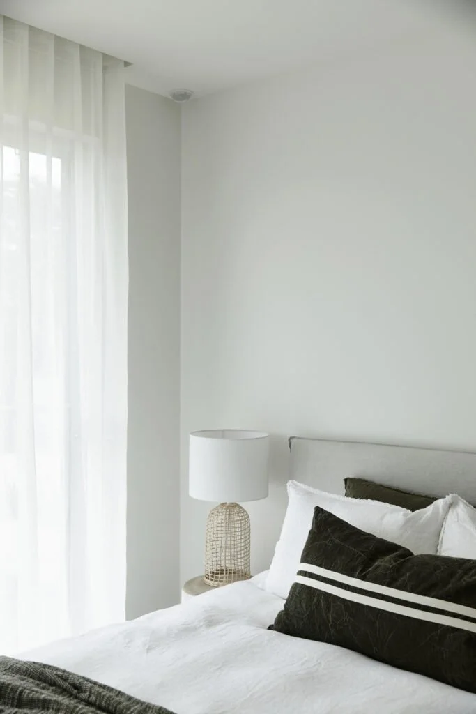 A bedroom featuring white walls, a bed with white bedding and black pillows, a gray headboard, a bedside table with a woven table lamp, and a window with sheer white curtains.