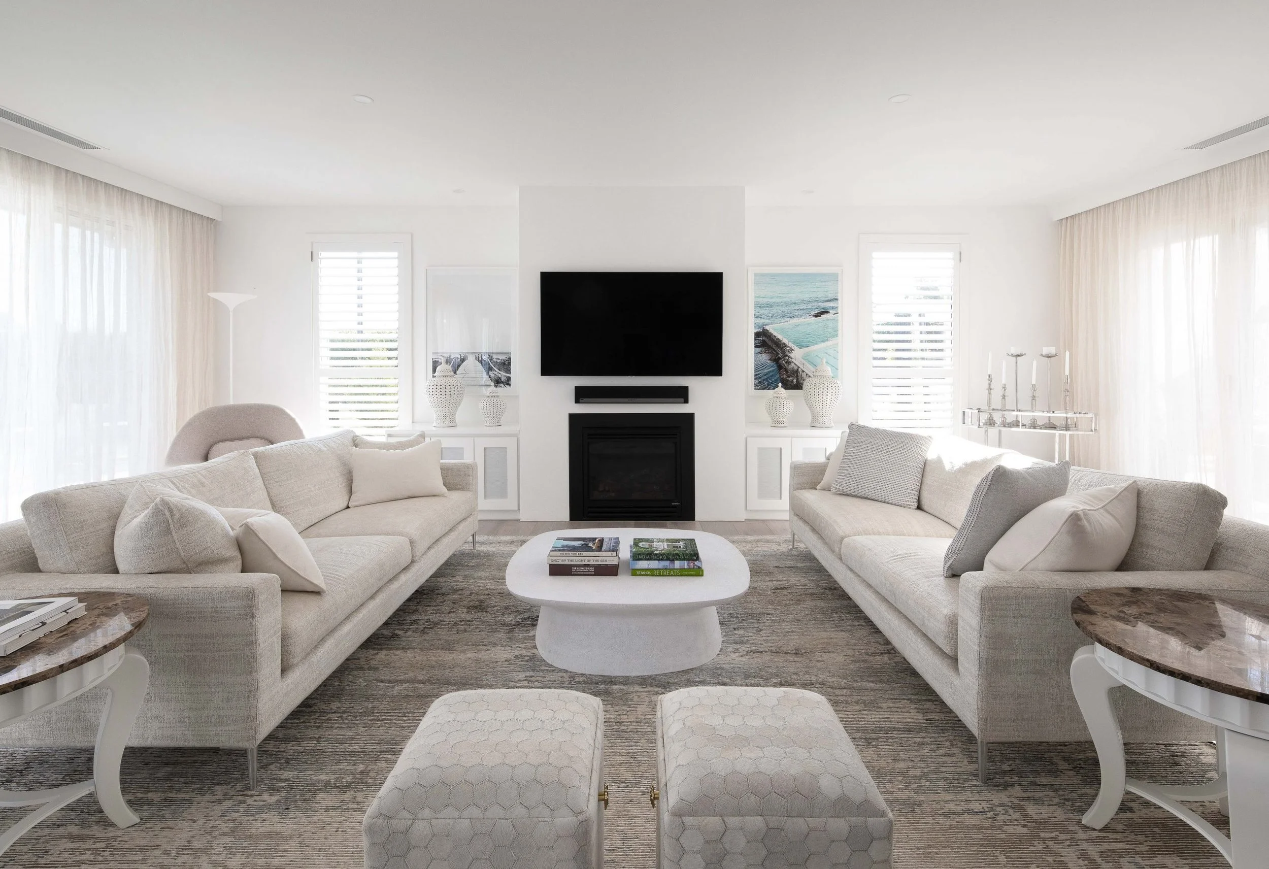 Bright living room with two cream-colored sofas, a white oval coffee table with books, framed ocean photos, a black fireplace, mounted TV, and large windows with light curtains.