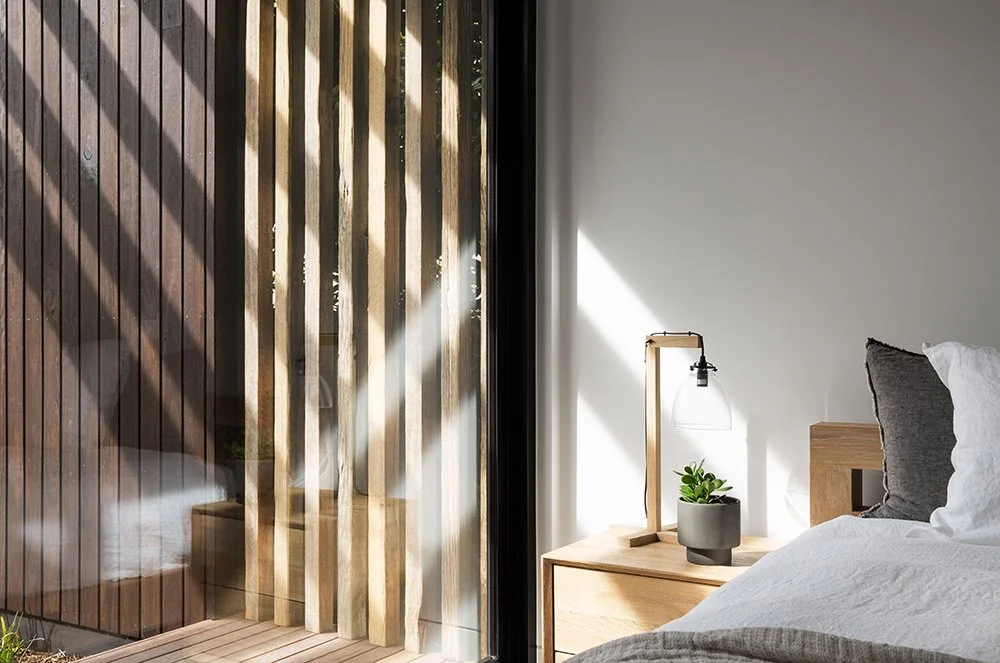 A bedroom with a wooden nightstand, a modern table lamp, and a small green plant, next to a bed with white and gray bedding. Outside, through a sliding glass door, is a wooden privacy fence and patterned shadows from sunlight.