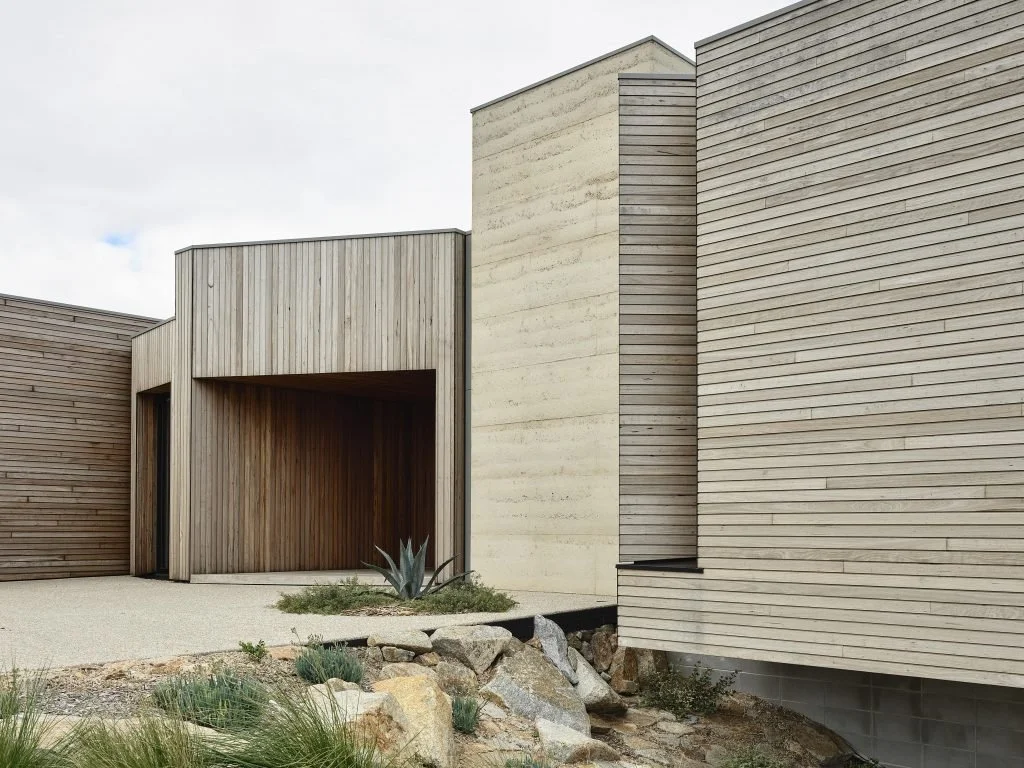 Modern house exterior with wooden and concrete siding, landscaped with rocks and plants.