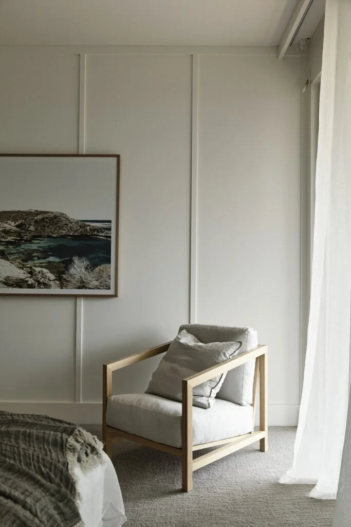 A cozy bedroom with a modern armchair made of light wood and white cushions, next to a bed with a textured blanket, a framed landscape photo on the wall, and a window with sheer white curtains.