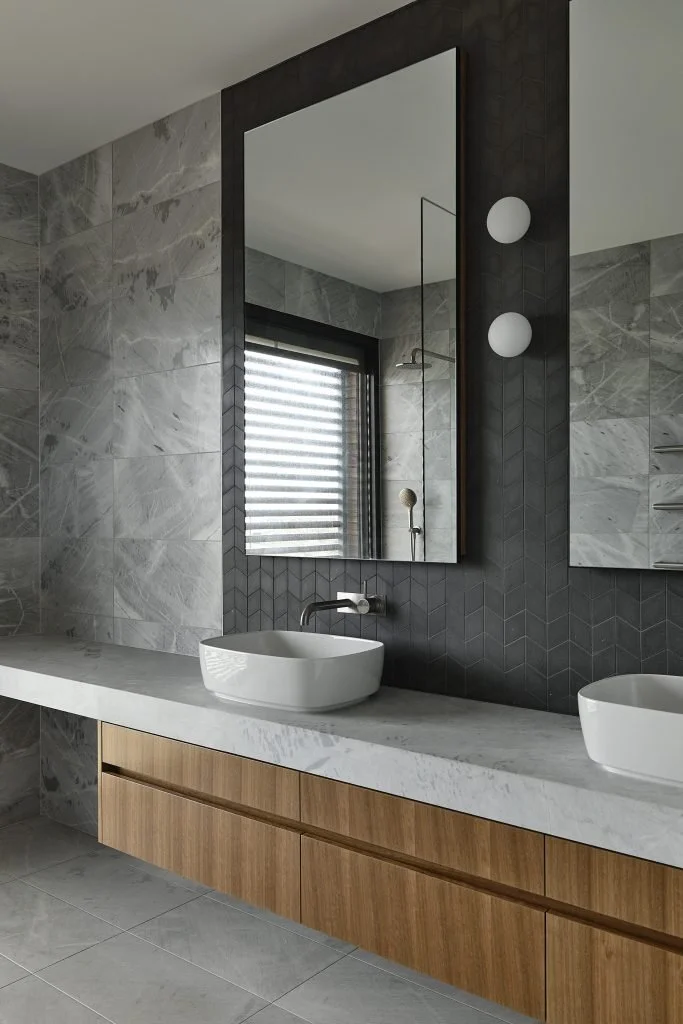 Modern bathroom with marble walls, a large mirror, a white sink vessel on a marble countertop, wooden cabinetry, and a window with horizontal blinds.