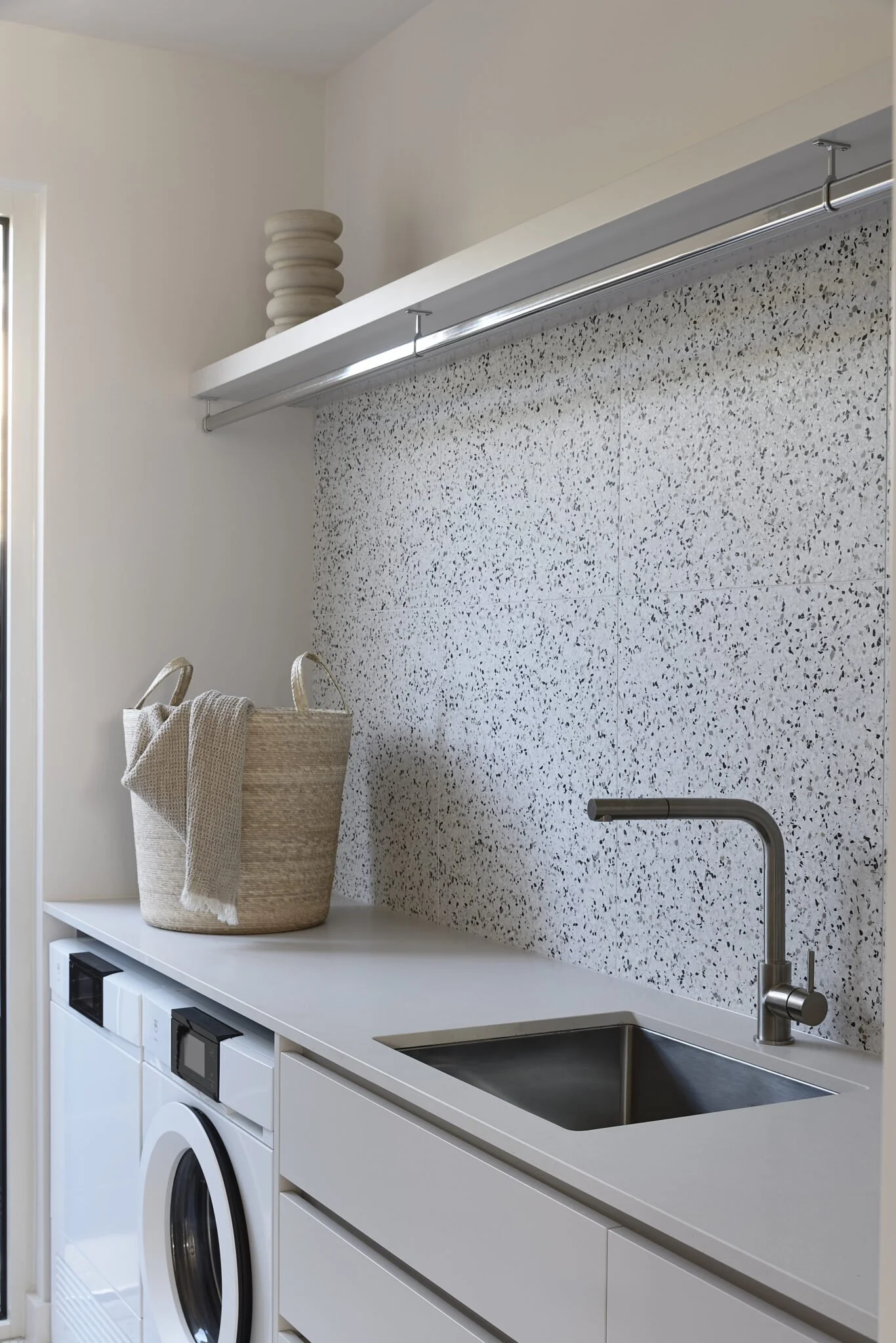 Laundry room with a white countertop, a small sink with a modern faucet, a washing machine, and a decorative vase on a shelf above. There is a woven laundry basket on the counter with some cloth inside.