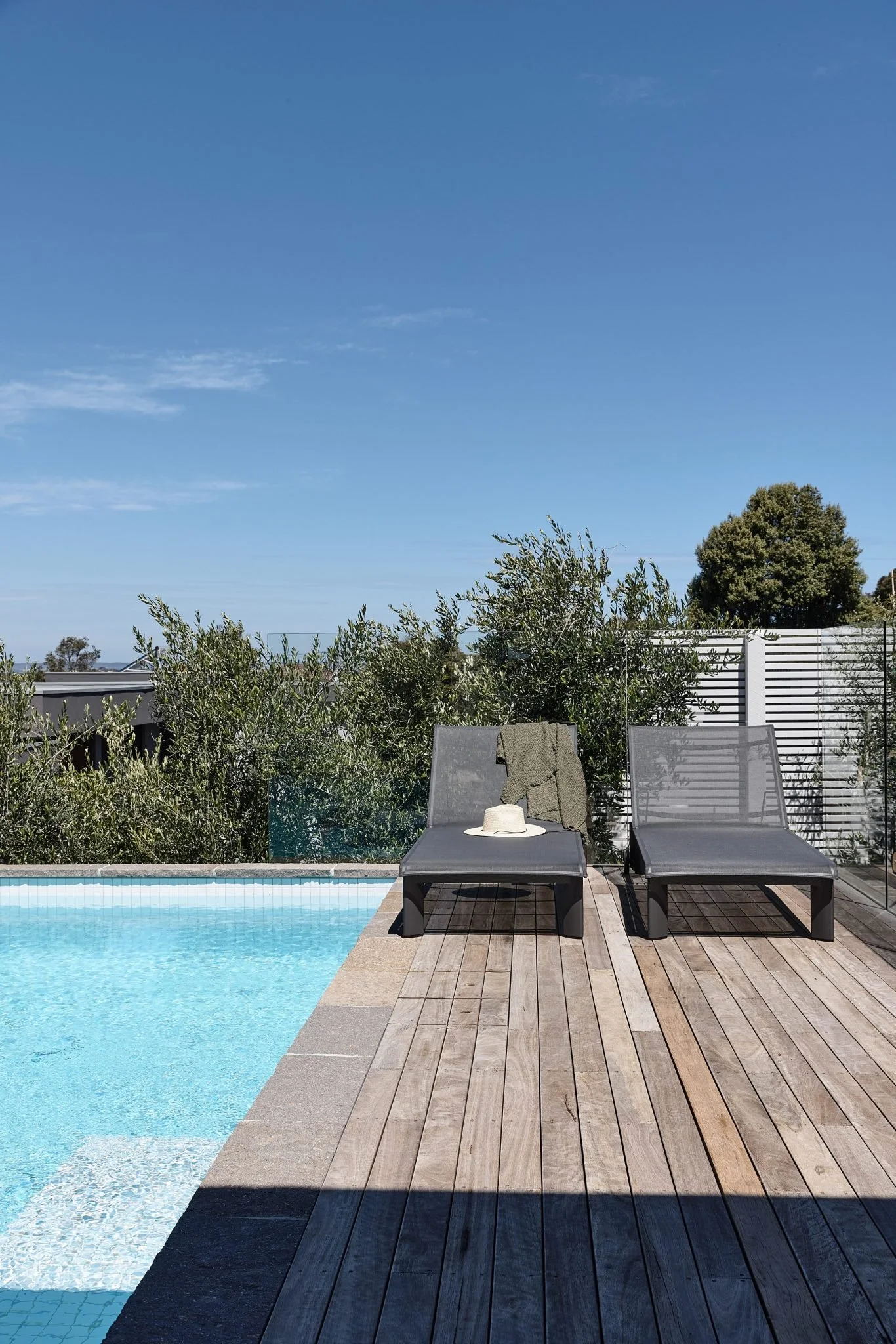 A backyard with a swimming pool, two black lounge chairs, a towel, a hat, and a wide blue sky.