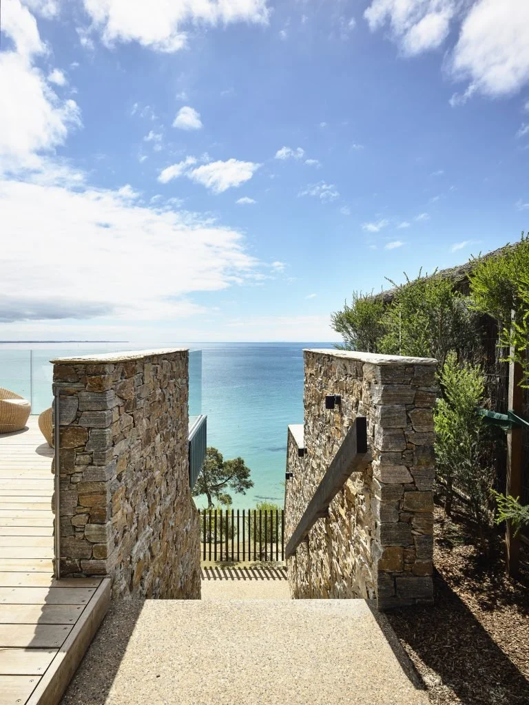 View of the ocean through a stone staircase with a wooden handrail, flanked by a deck on the left and greenery on the right, under a partly cloudy sky.