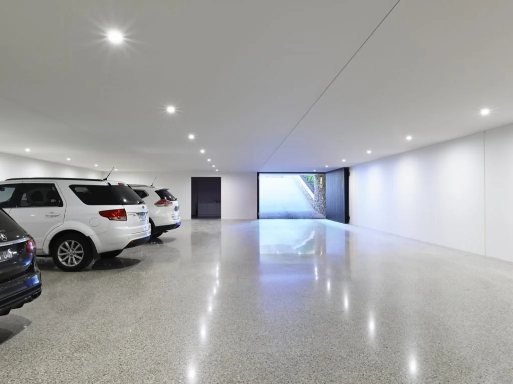 A clean, empty underground parking garage with a few parked cars along the left side and open space towards a large entrance or exit at the back, illuminated by ceiling lights.