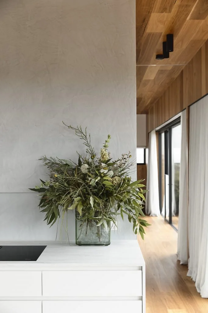 A bouquet of greenery in a glass vase on a white dresser in a modern room with wooden accents and large windows.