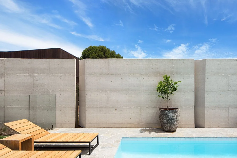 Modern outdoor pool area with a concrete wall, potted plant, lounge chairs, and blue sky.