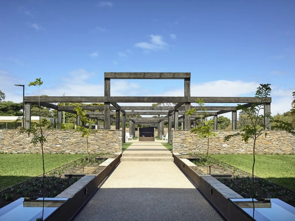 A modern outdoor garden with a stone wall, wooden pergola structure, and a paved walkway leading to a building with a black facade.