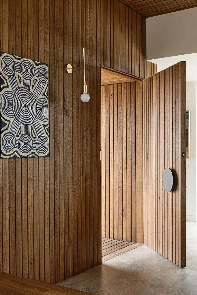 A wooden interior wall with a sliding wooden door partially open, exposing a small entryway with wooden flooring, and a modern geometric wall art piece on the left.