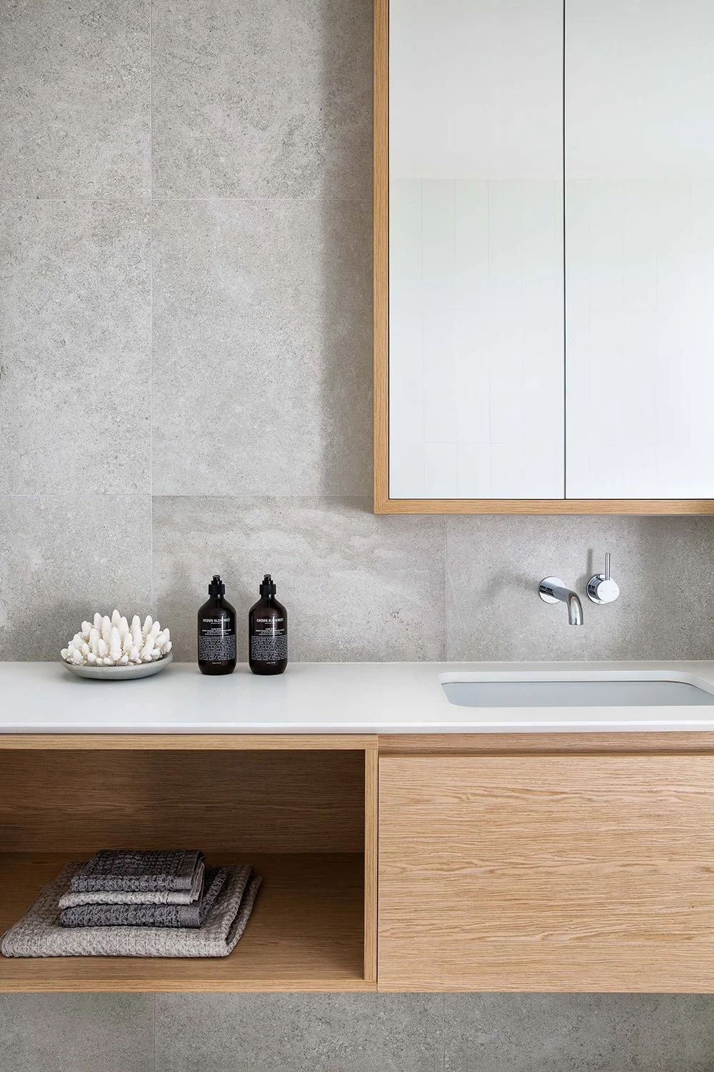 Modern bathroom vanity with a white countertop, two black bottles, a decorative white coral, and a mirrored cabinet above, against a beige tiled wall.