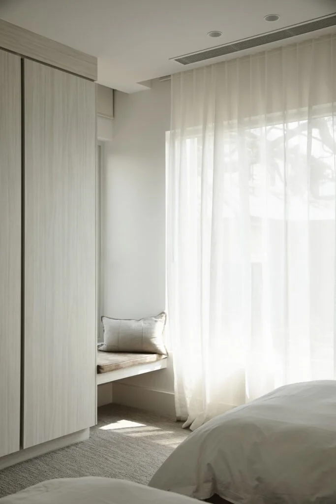 Bright bedroom corner with white curtains, a window seat with a pillow, built-in wardrobe, and a bed with white linens.