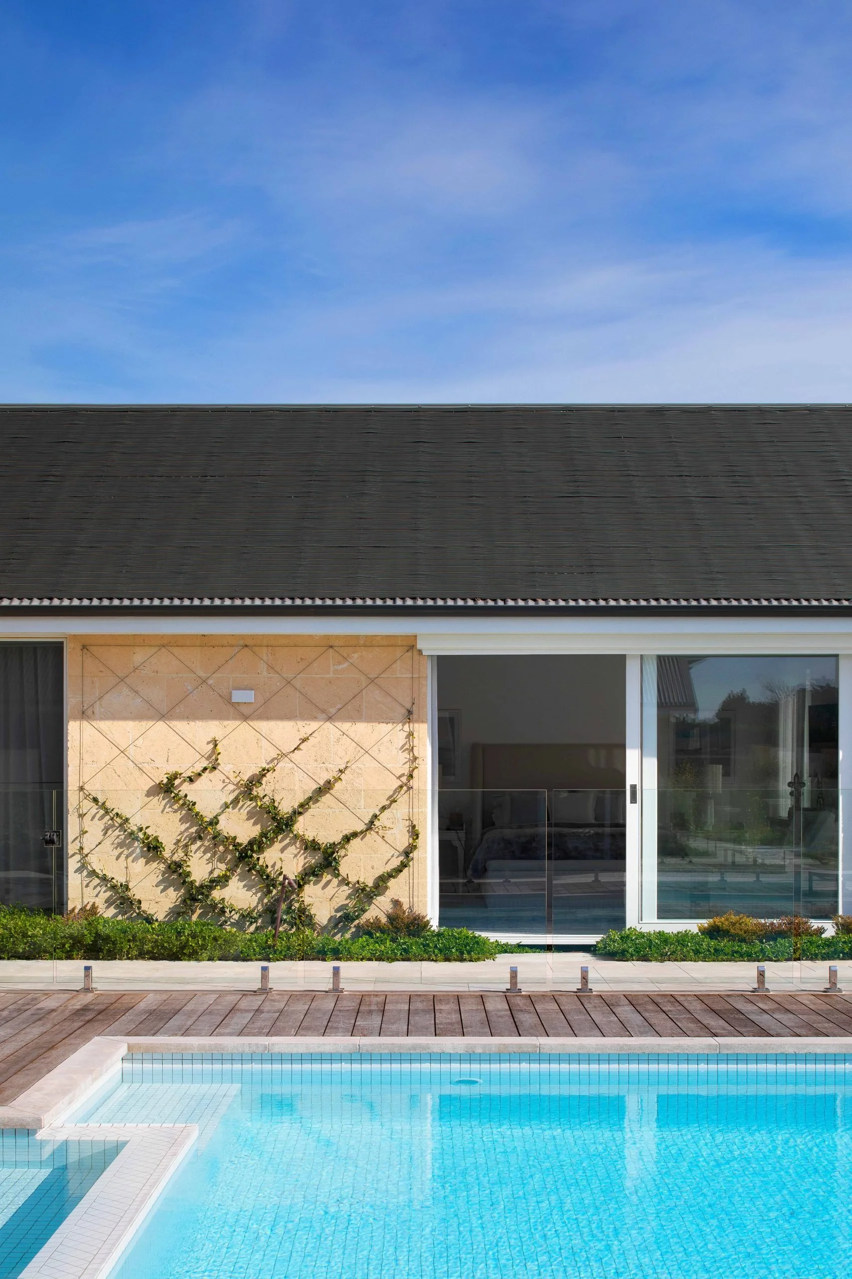 Modern house with a dark roof, glass sliding door, and a small garden with climbing plants, next to a swimming pool with a wooden deck.