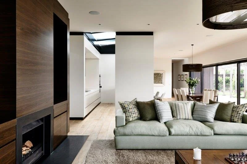 Modern living room with light-colored sofa and decorative pillows, dark wood accent wall with fireplace, open dining area with large window and sliding glass door, potted plant, and minimalistic decor.