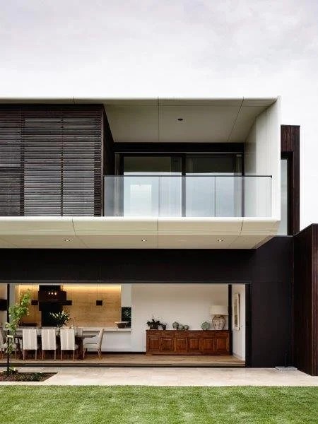 Modern two-story house with a balcony, large glass windows, and an open-plan backyard.