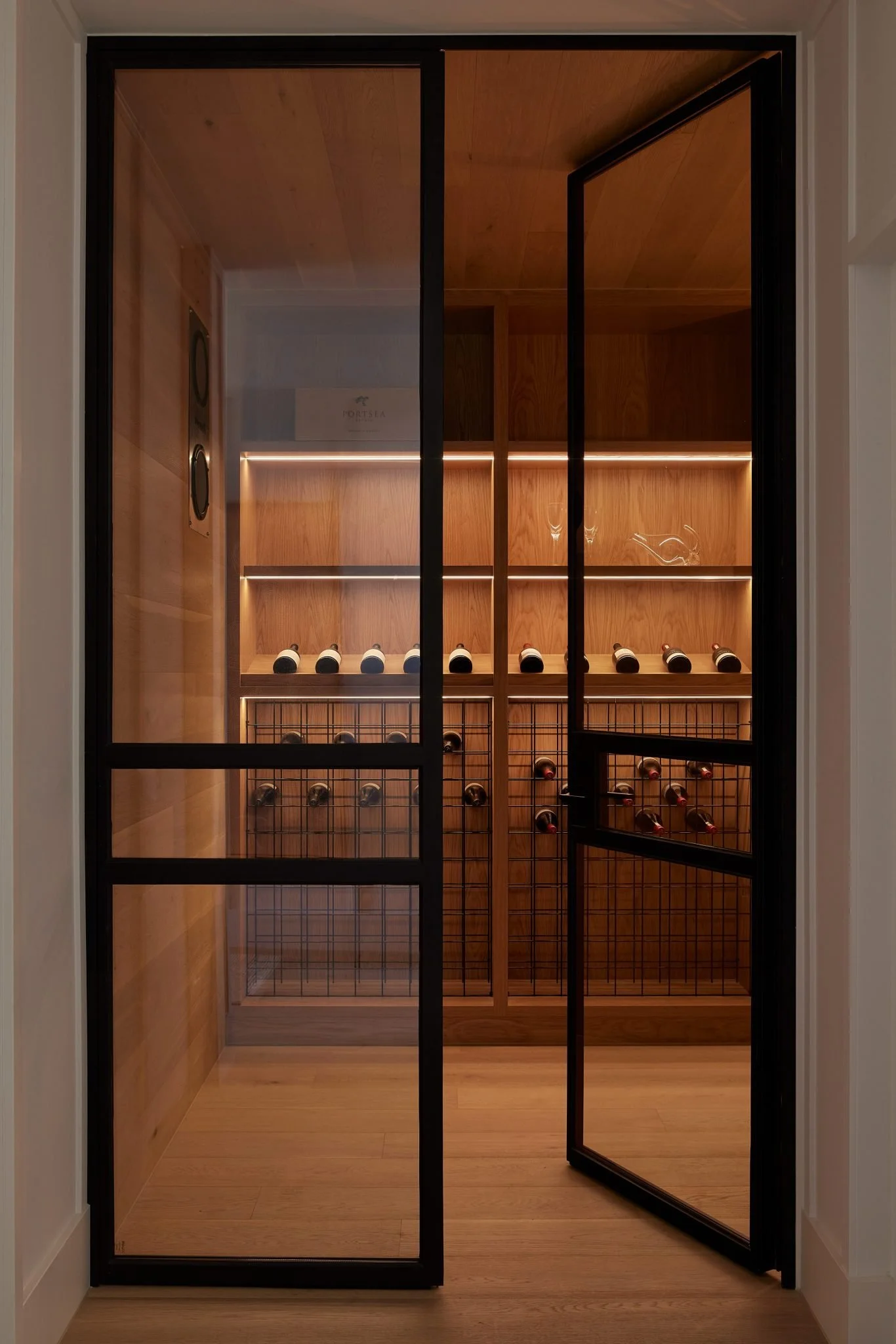 A wine cellar with wooden shelves and wine bottles stored on racks, viewed through a black-framed glass door.
