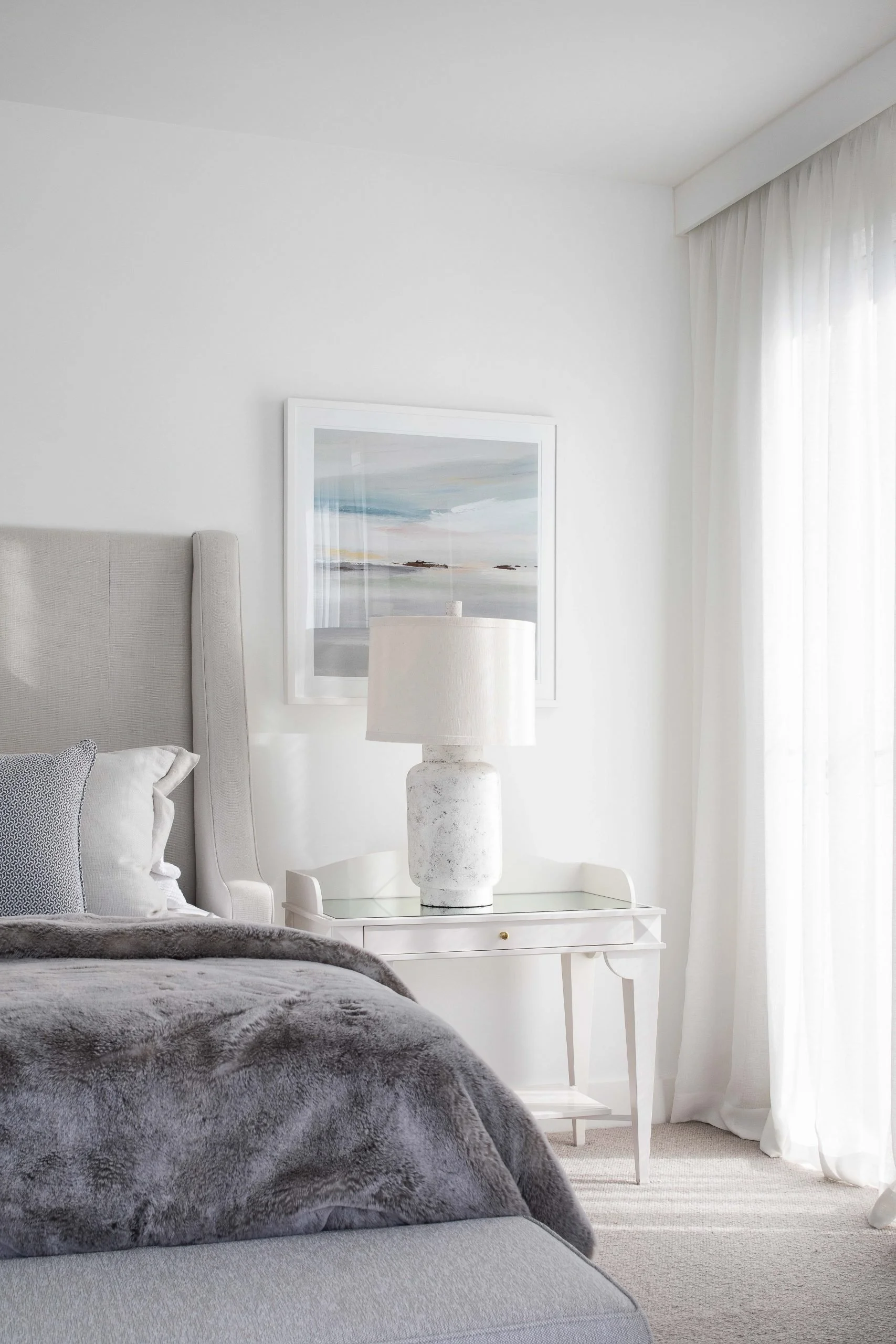 A bedroom with a gray upholstered bed, a white nightstand with a large marble lamp, a framed artwork of a seascape above the nightstand, white curtains, and a window letting in natural light.