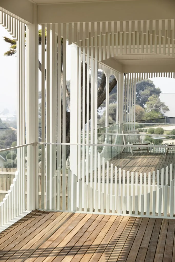 Modern outdoor balcony with wooden flooring and white vertical slat railing, overlooking trees and landscape.