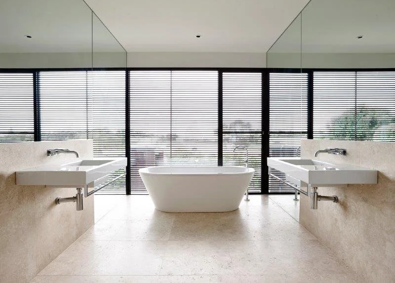 Modern bathroom with a freestanding bathtub centered in front of floor-to-ceiling windows with black horizontal blinds, flanked by two wall-mounted sinks.