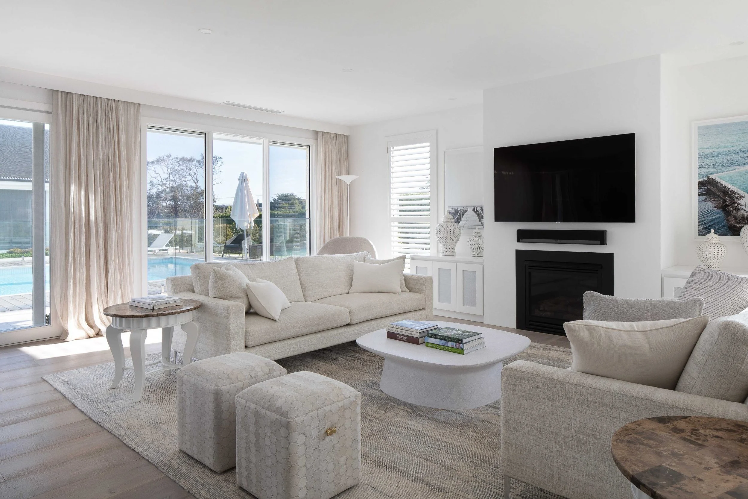 Bright living room with white sofas, a TV above a fireplace, a sliding glass door leading to a pool area, and large windows with blinds and curtains.