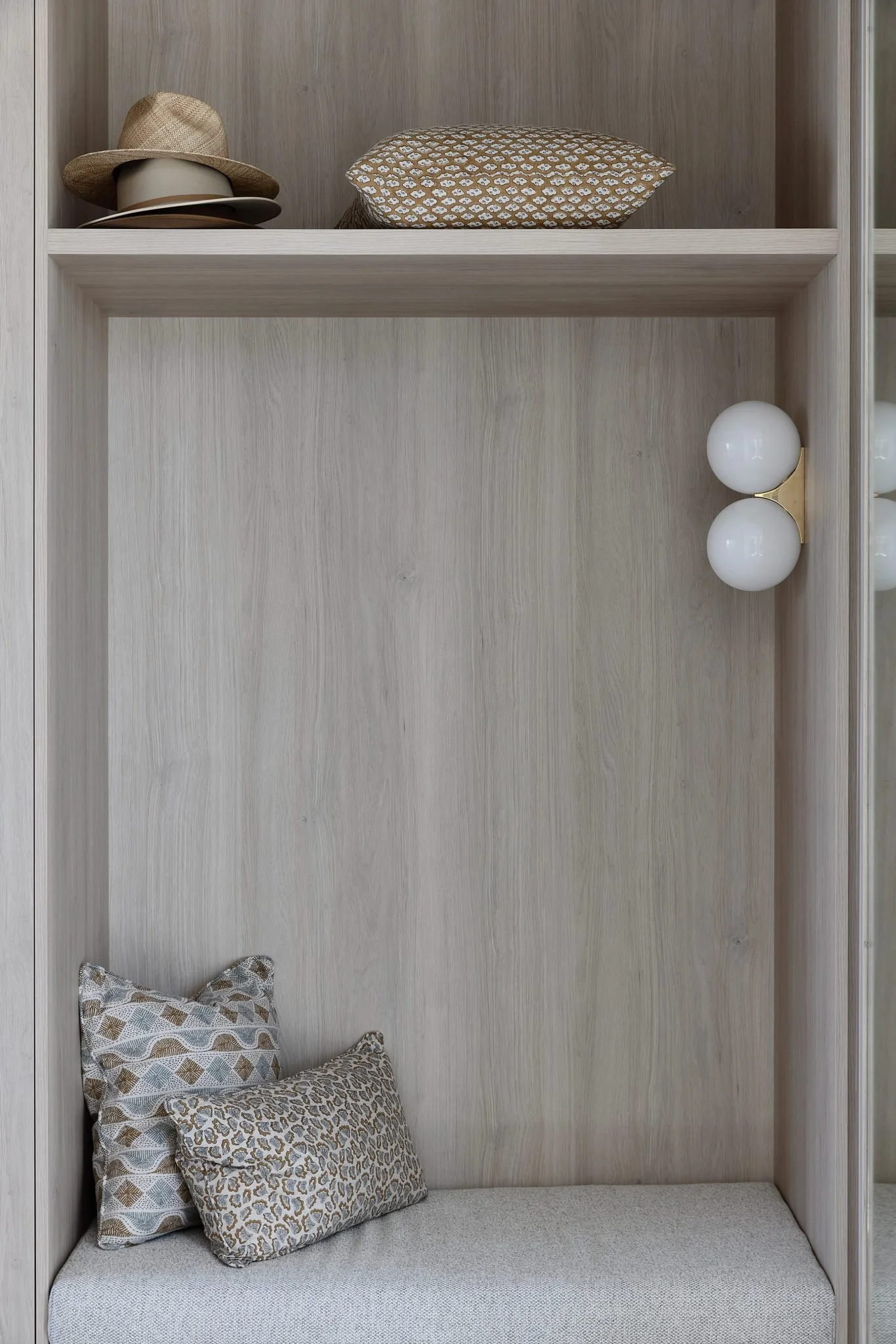 A built-in storage nook with a light wood interior, featuring a small bench with two patterned pillows, a modern wall sconce with two white globe bulbs, and a shelf holding hats and a patterned pillow.