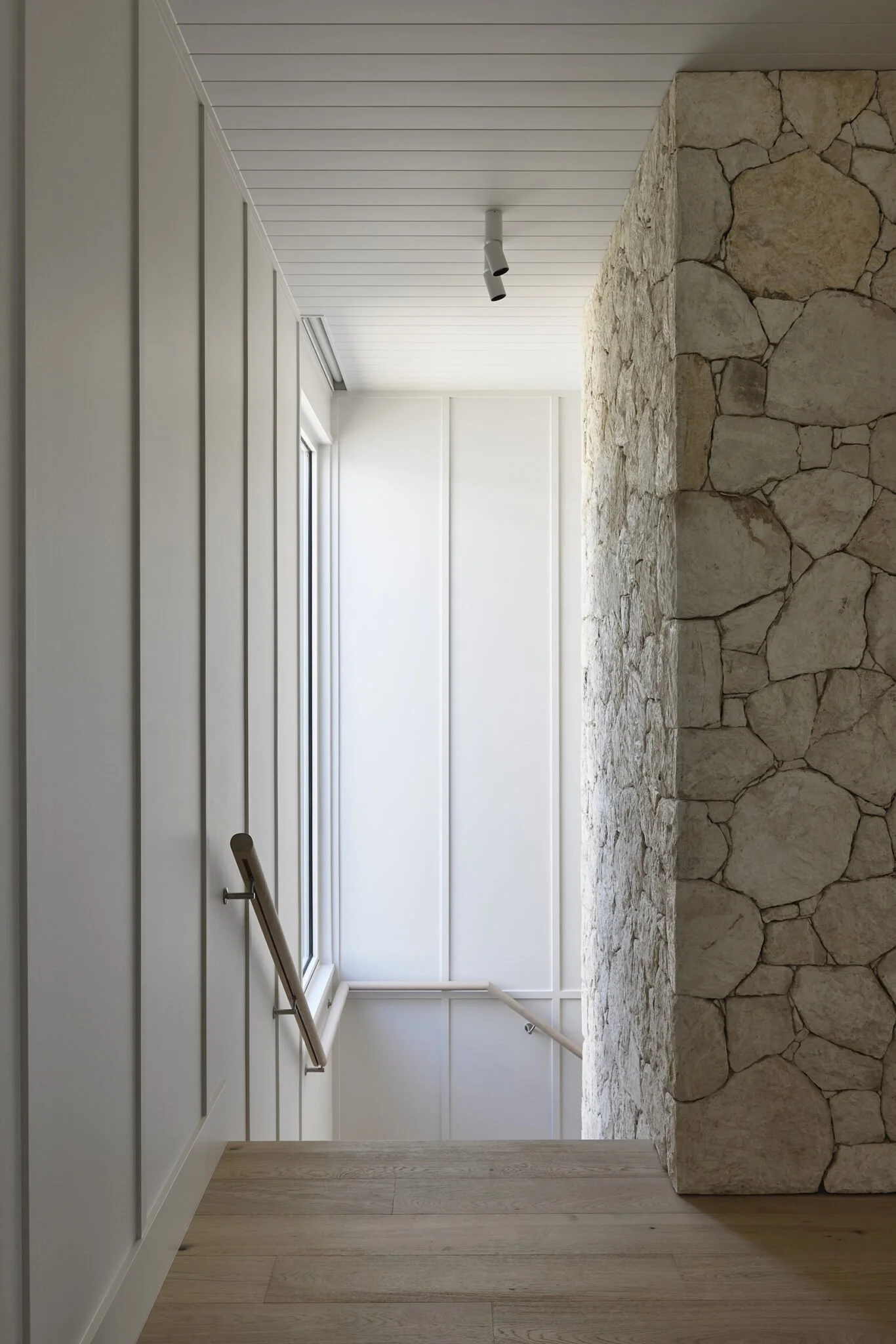 Interior view of a staircase landing with white paneling, wood flooring, a stone wall, and a window.
