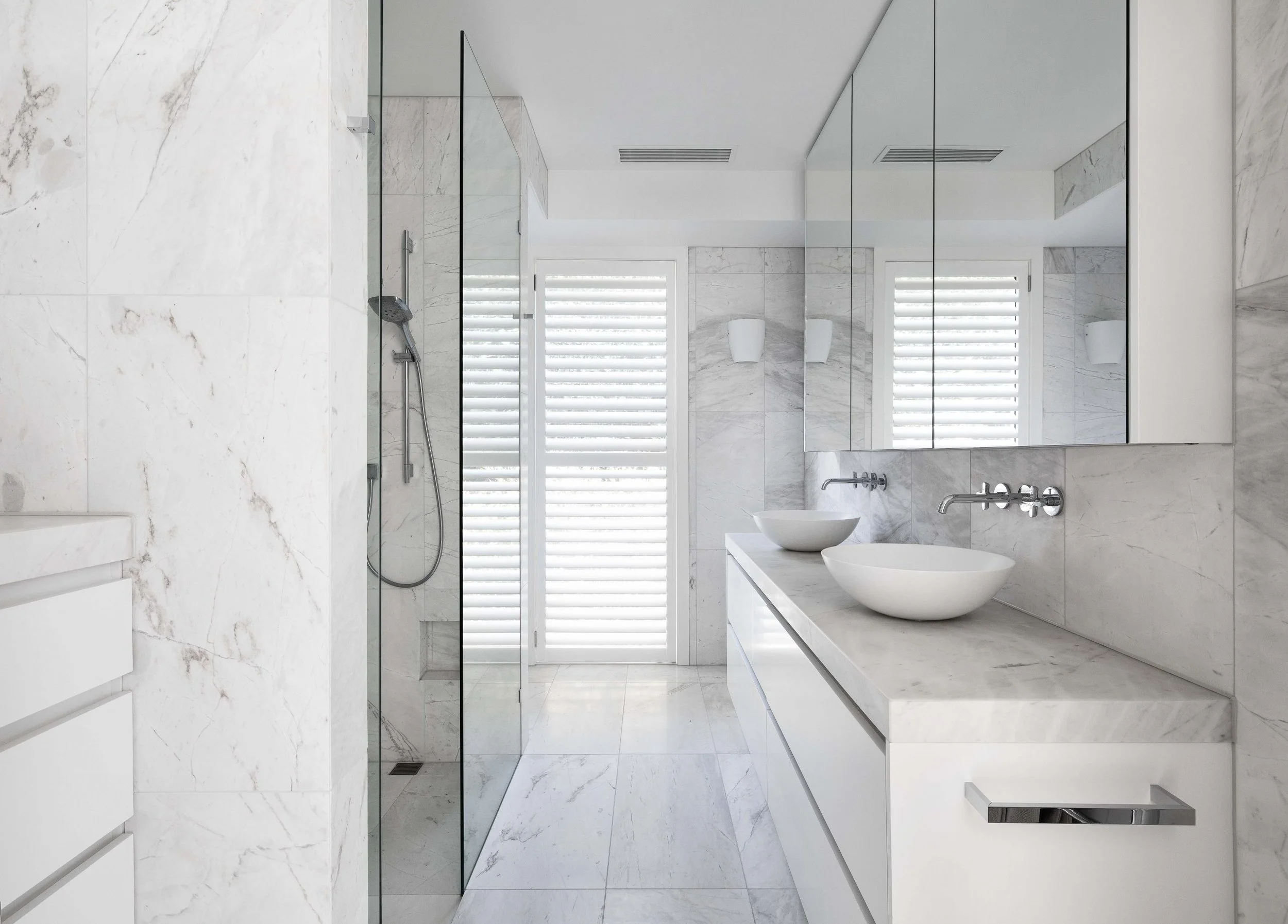 Modern white bathroom with marble walls and floor, two vessel sinks on a marble vanity, large mirror, a showerwith glass door and a window with white shutters.