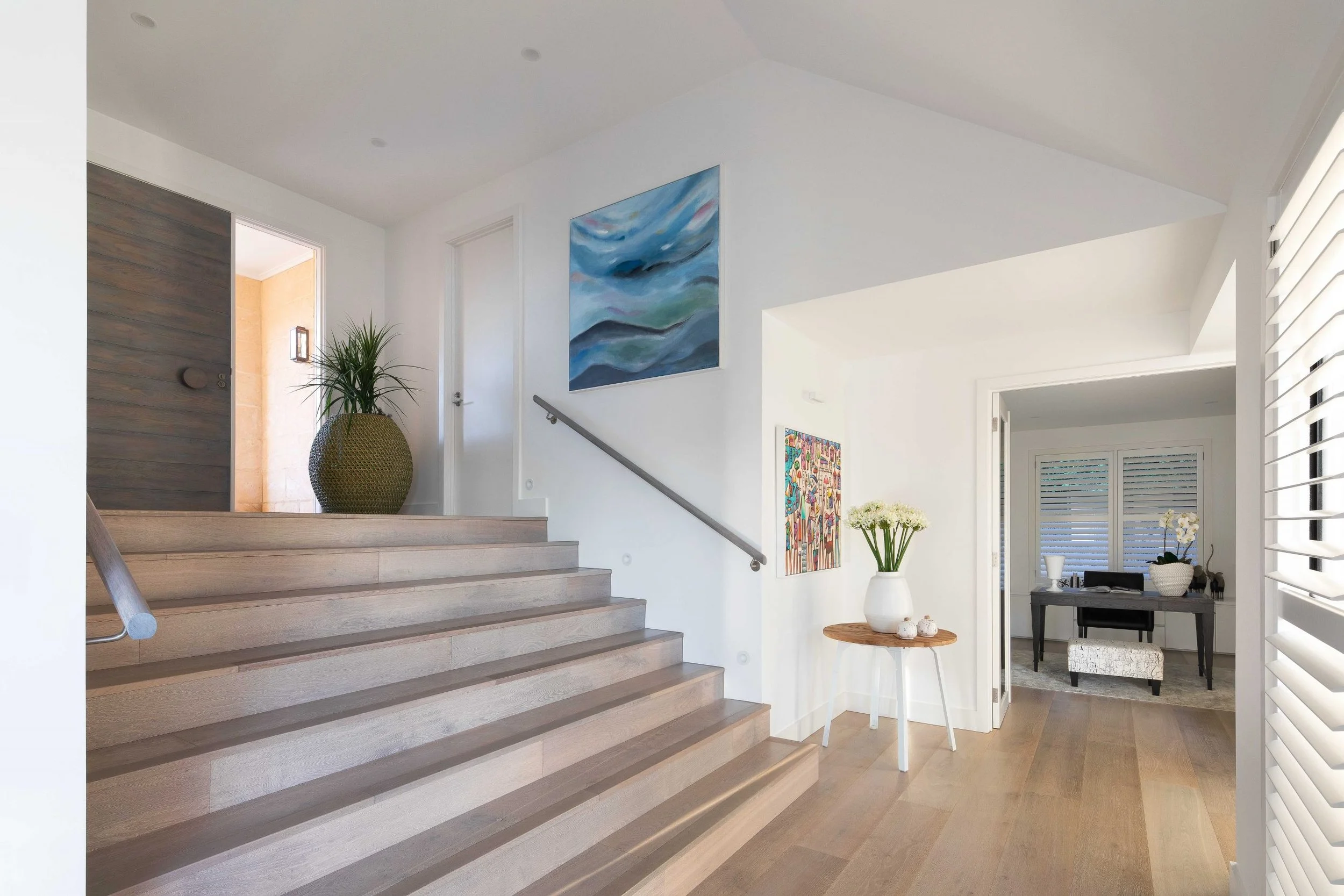 Bright hallway with wooden stairs, white walls, colorful paintings, potted plants, and an adjoining room with a desk and window.