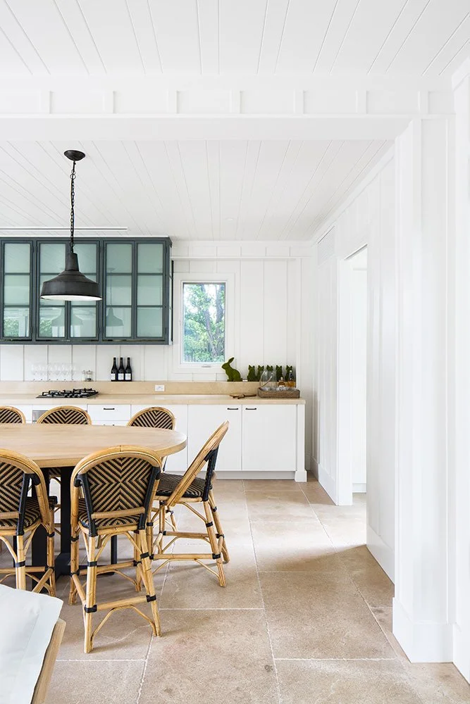 A bright kitchen with white Paneled walls and ceiling, a wooden dining table with rattan chairs, a window showing trees outside, and a countertop with plants and bottles.