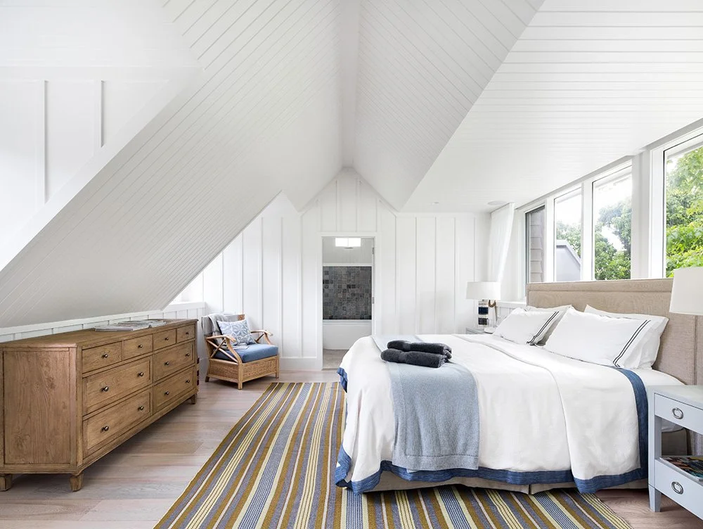 Bright bedroom with sloped white ceiling, large windows, beige headboard, white bedding with blue trim, wooden dresser, armchair, and striped rug.