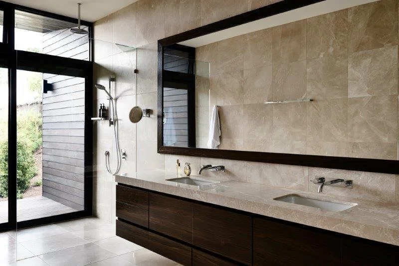 Modern bathroom with double sinks, large mirror, and a walk-in shower with glass door and black accents. Light-colored tiled walls and floor, and a towel hanging on the wall.