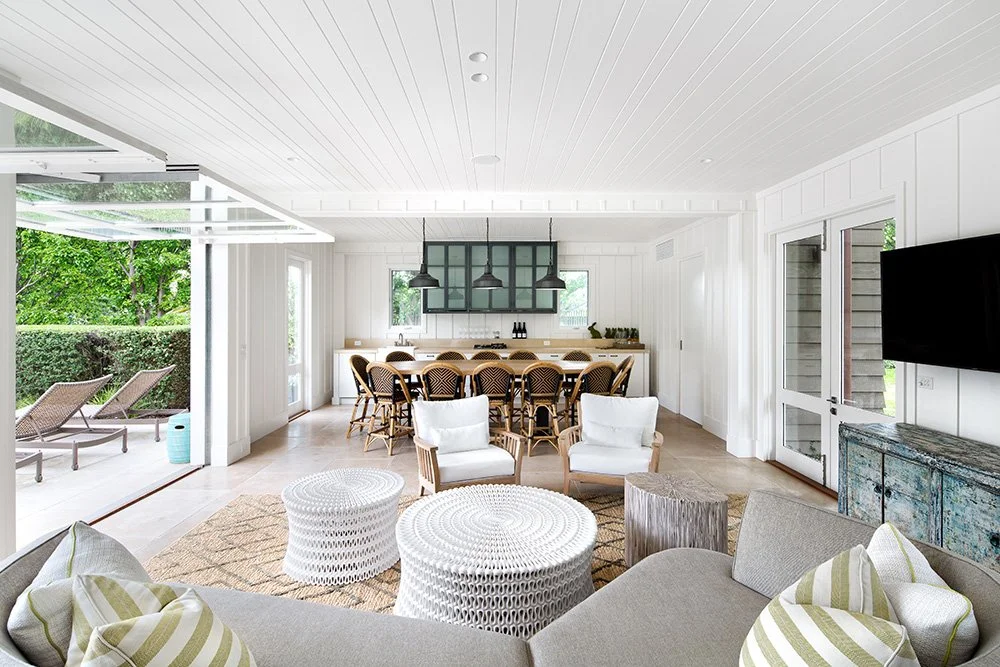 Modern bright living room with white paneled walls, a large flat-screen TV, wicker armchairs with white cushions, a beige sofa with striped pillows, two round white woven coffee tables, and sliding glass doors leading to an outdoor patio with lounge