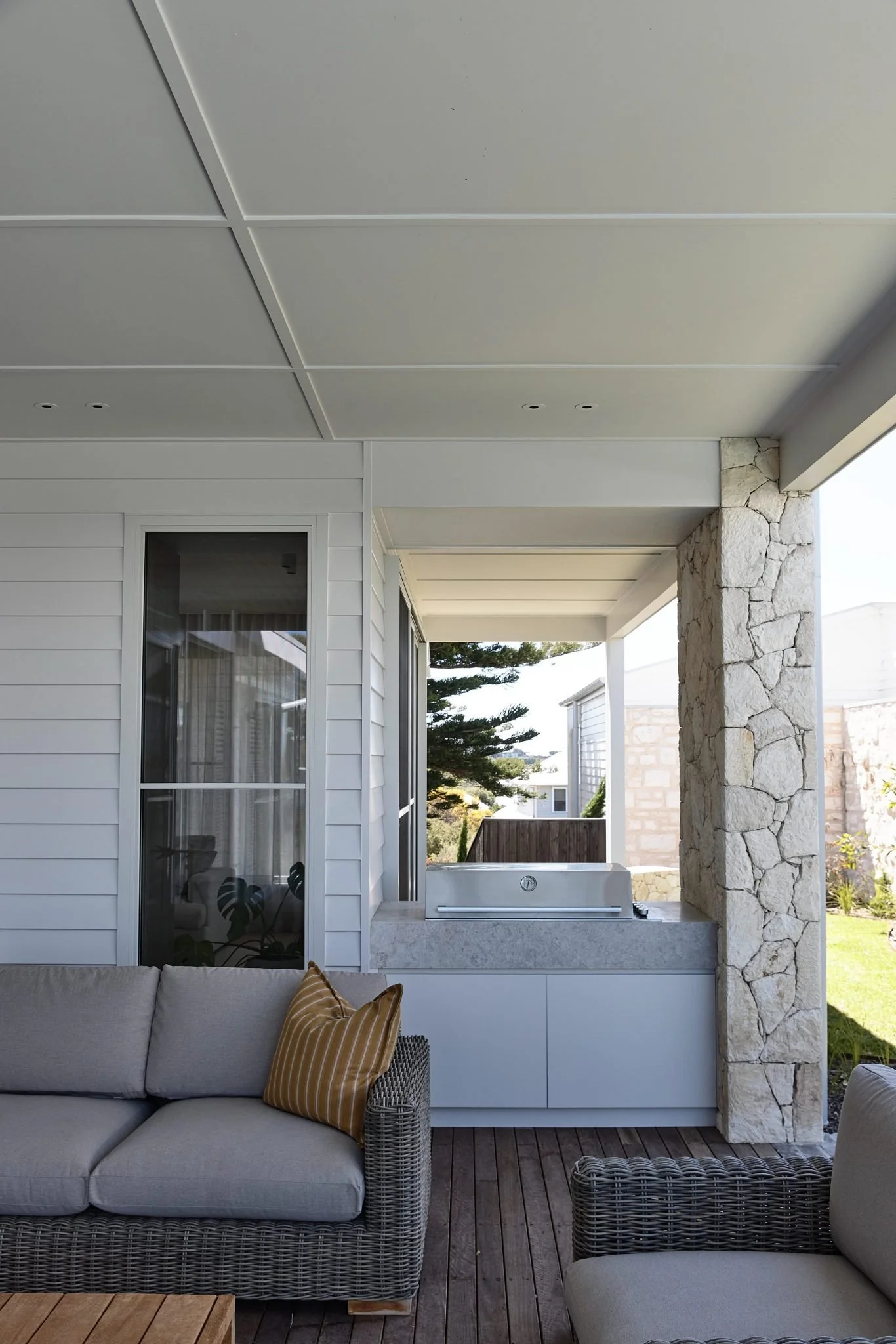 Outdoor patio with wicker sofa, beige cushion, yellow striped pillow, and hot tub beside stone pillar, with trees and part of house visible in the background.
