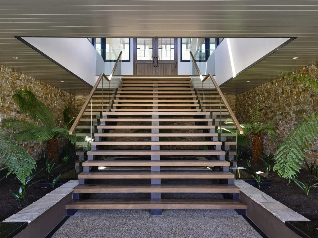 Indoor staircase with wooden steps and glass railings, flanked by potted ferns and stone walls, leading up to a double door with glass panes.
