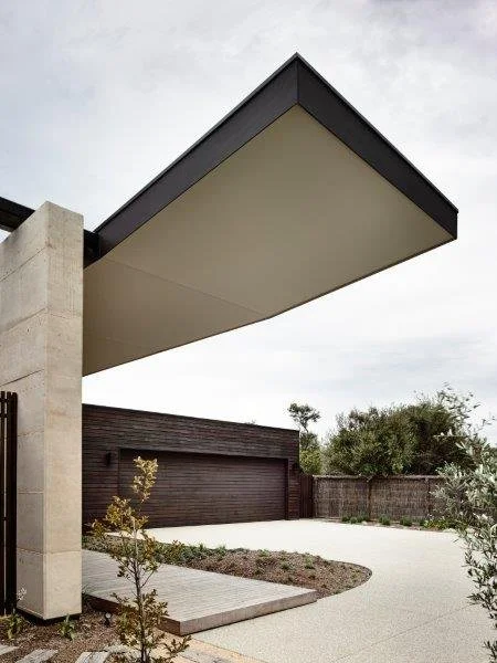 Modern house with an overhanging flat roof, wooden exterior, driveway, and surrounding landscaping.