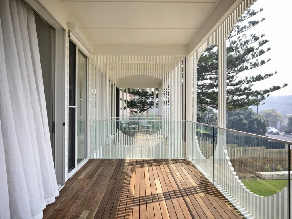 A balcony with wooden flooring and a modern glass railing, featuring vertical white slats and a view of trees and houses in the distance.