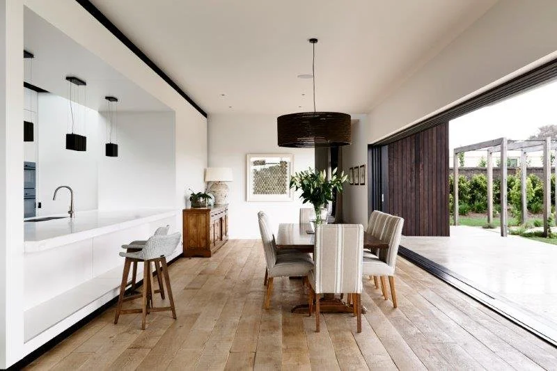 Open concept dining area with a wooden table, six upholstered chairs, a large black pendant light, a sliding glass door leading outside, a kitchen with a white island, Barstools, and minimal decor.
