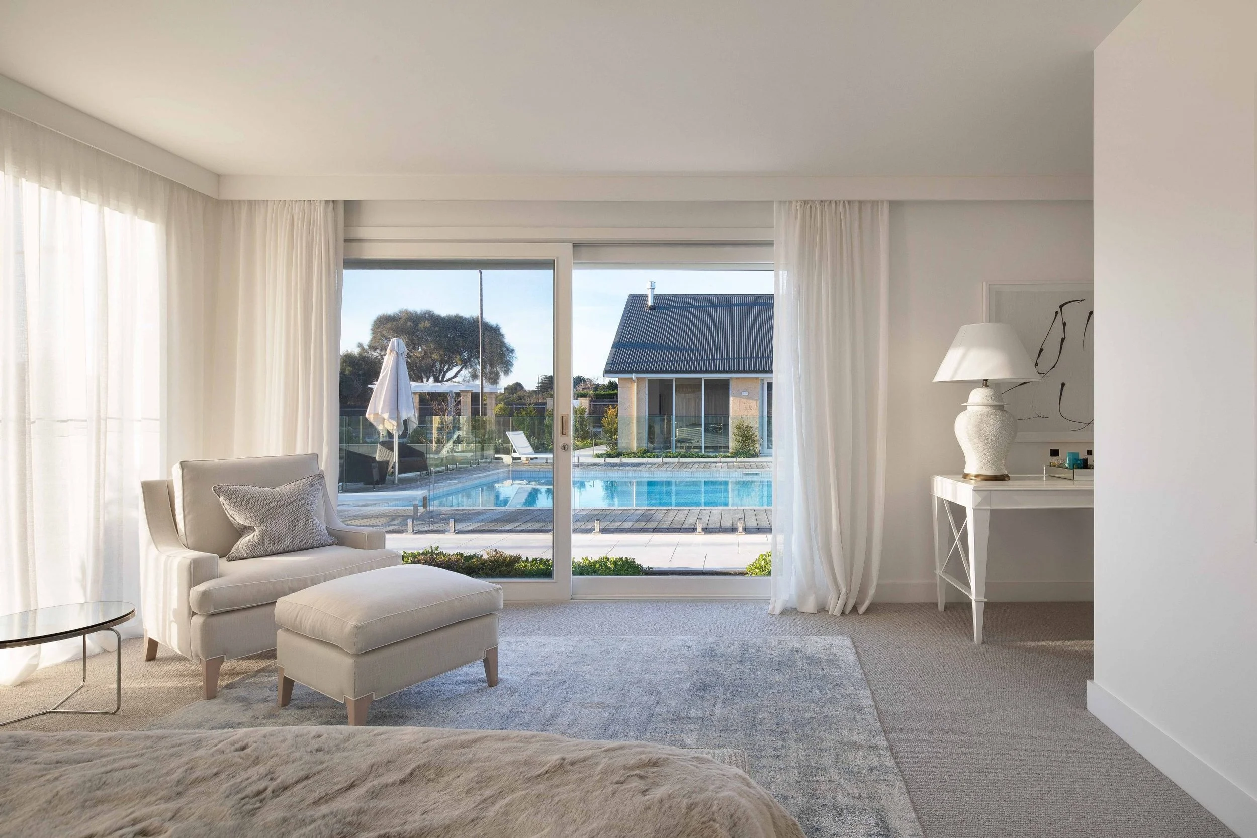 Bright bedroom with large glass sliding door revealing a swimming pool outdoors, with white curtains, a beige armchair with cushion, a matching ottoman, a white side table, a decorative lamp, and modern artwork.