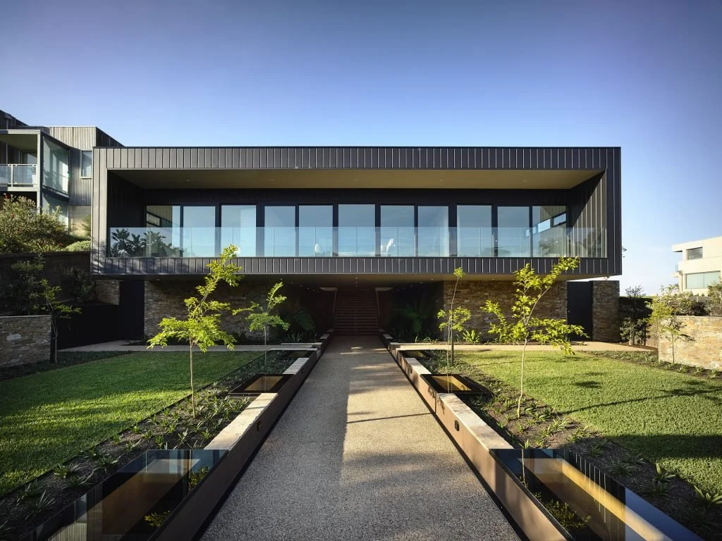 Modern two-story house with large glass windows and metal siding, surrounded by a landscaped garden with young trees and planters.
