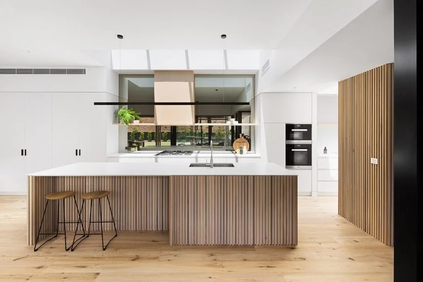 Modern kitchen with white cabinets, wooden accents, and a large kitchen island with a white countertop. Two bar stools are placed at the island. There are built-in appliances and a window with a view of greenery outside.