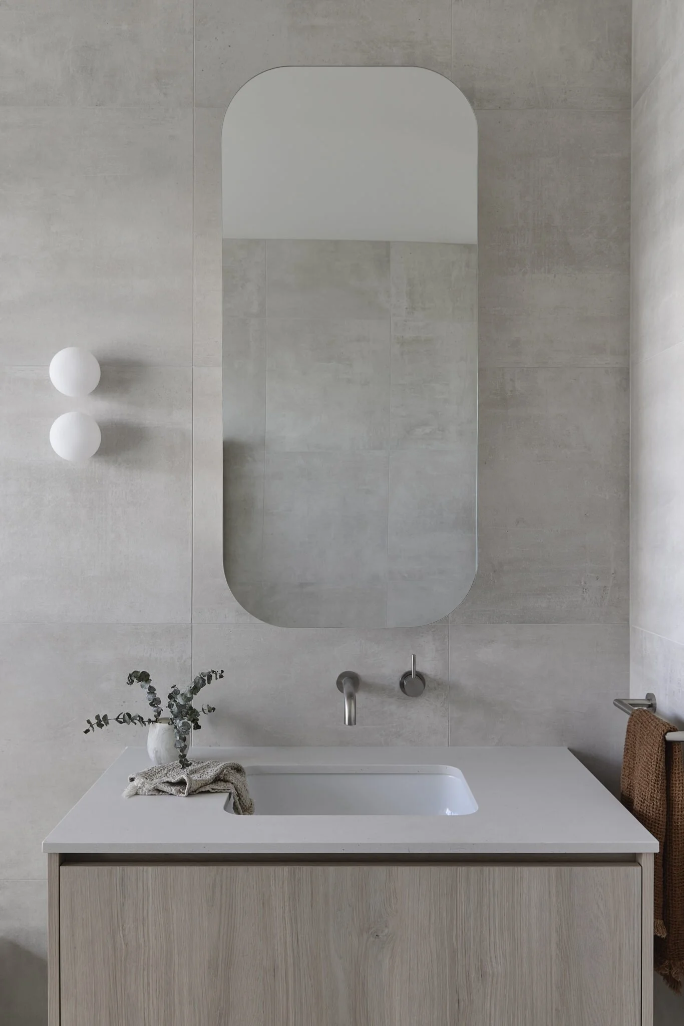 Modern bathroom with a light wooden vanity, a rectangular sink, a large vertical mirror, a wall-mounted faucet, eucalyptus in a white vase, a folded cloth, and a brown towel on a towel rack, all in neutral tones.