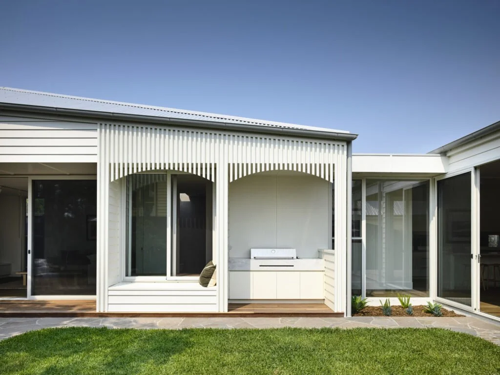 Modern house with white siding and large glass sliding doors; small deck area with built-in grill in backyard with green grass and a blue sky.