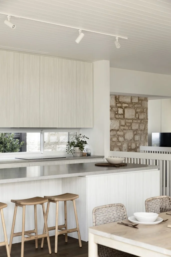 Modern kitchen with white cabinetry, a long countertop, a window with a plant, and three wooden bar stools.
