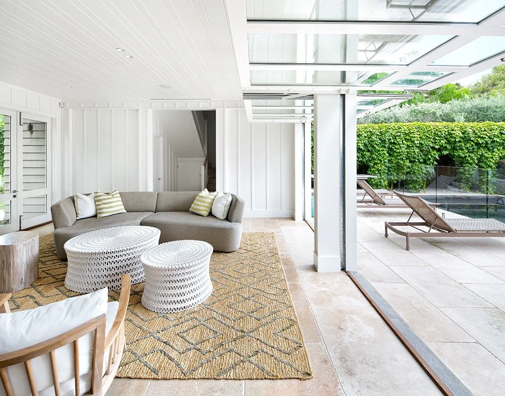 Interior view of a modern living room opening to an outdoor patio with lounge chairs, greenery, and glass railing.