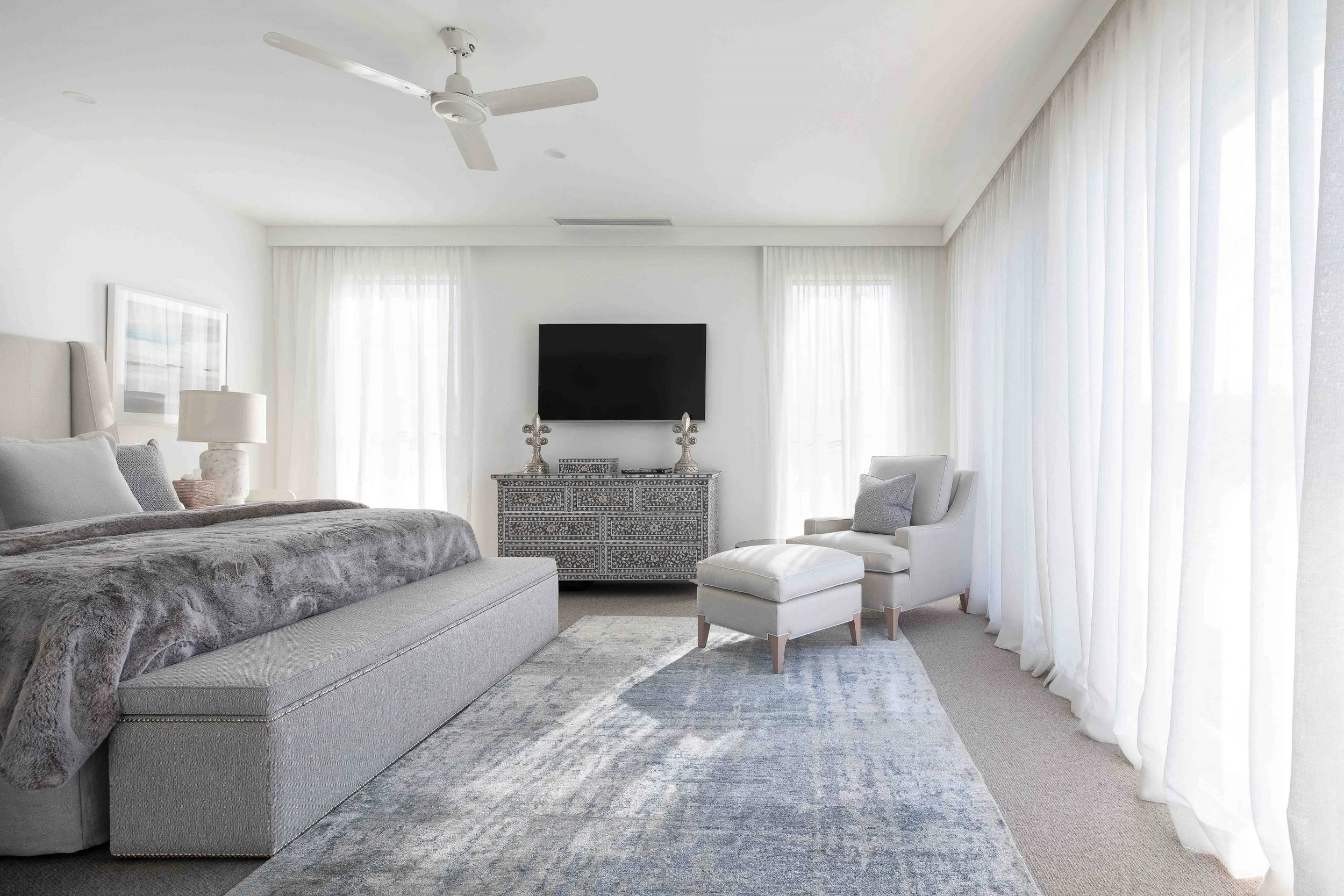 Bright bedroom with white walls, ceiling fan, large windows with sheer white curtains, a bed with gray blankets, a white upholstered armchair with a gray pillow, a rug, a patterned dresser with decorative items, and a mounted flat-screen TV.