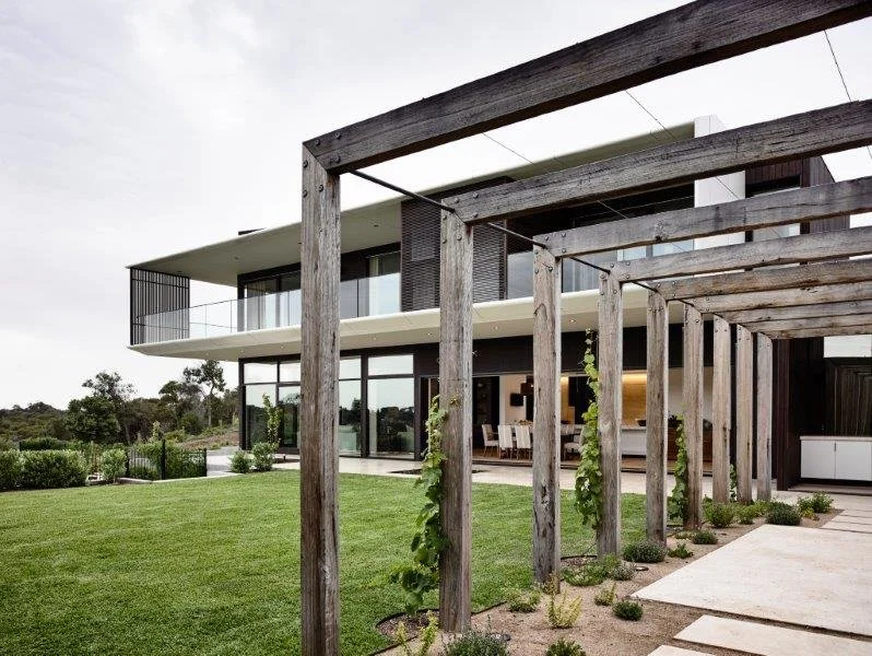 Modern house with glass walls and a balcony, wooden pergola with plants, green lawn, and outdoor seating area.