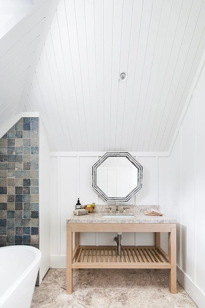 Bathroom with white paneled walls and ceiling, a light wood vanity with a stone countertop, a decorative octagonal mirror, a small sink, and a tiled wall with multi-colored square tiles.